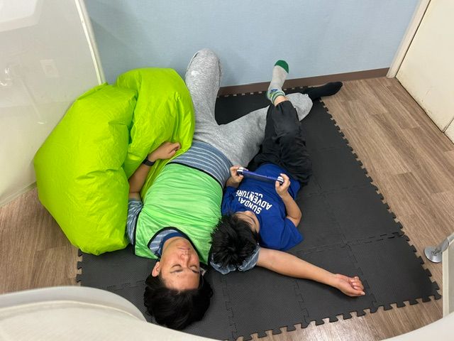 マットの上でのんびり過ごす学生スタッフとこども